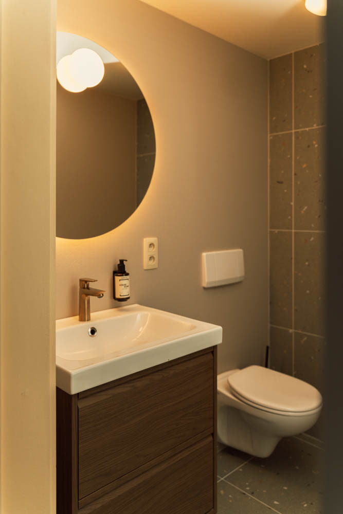 Modern en-suite hotel bathroom with round backlit mirror, vanity basin, and brass fittings