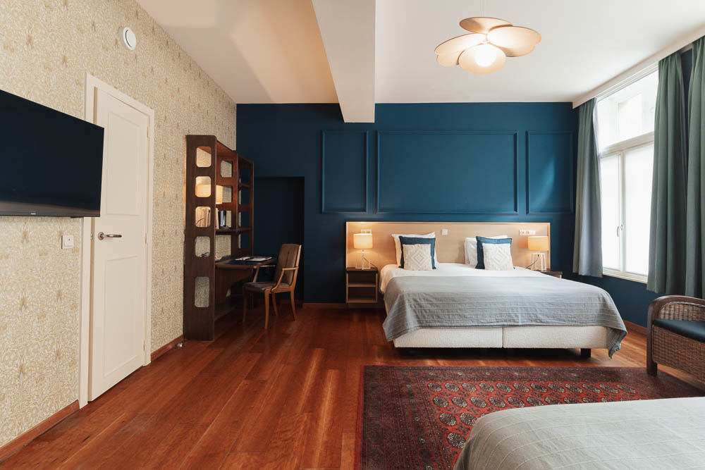 Spacious blue-painted hotel room with double bed, smart TV, bookshelf, and daylight