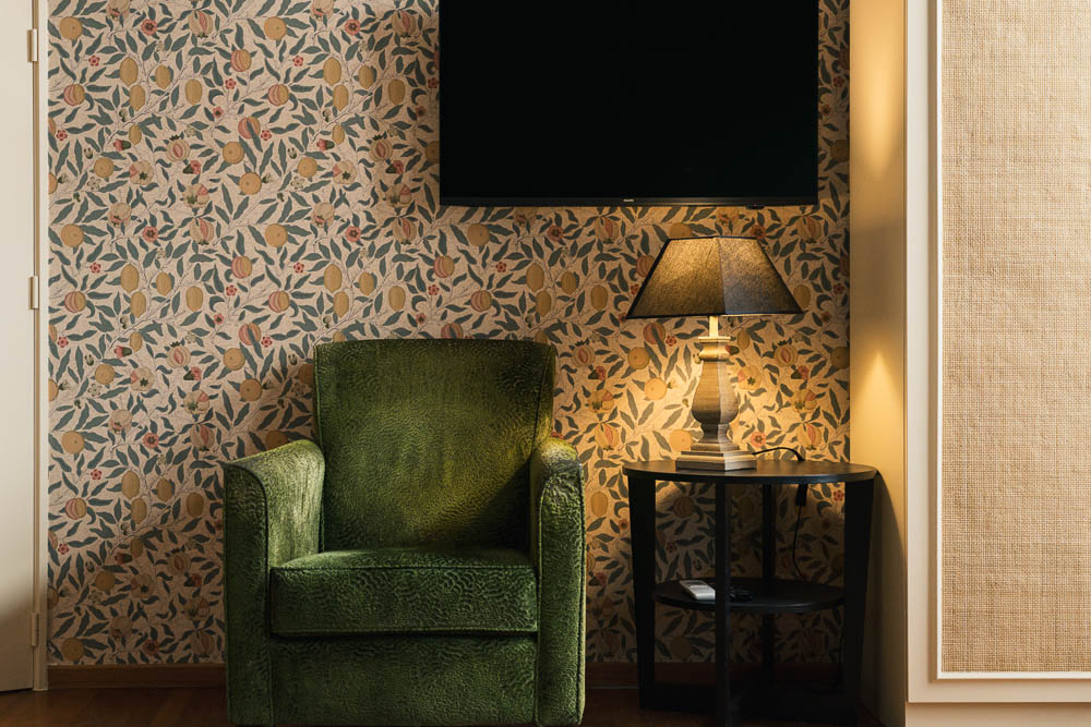 Cosy hotel room corner with green velvet armchair, botanical wallpaper, and side lamp
