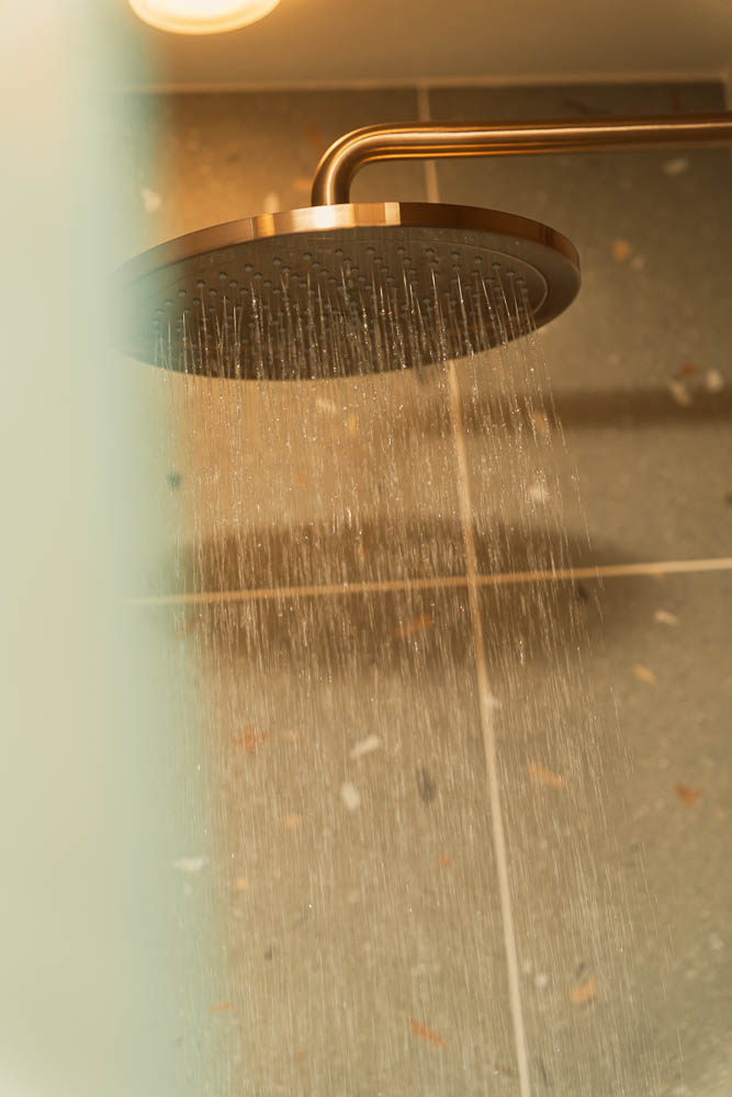 Overhead brass rain shower head running in full flow against warm tiles